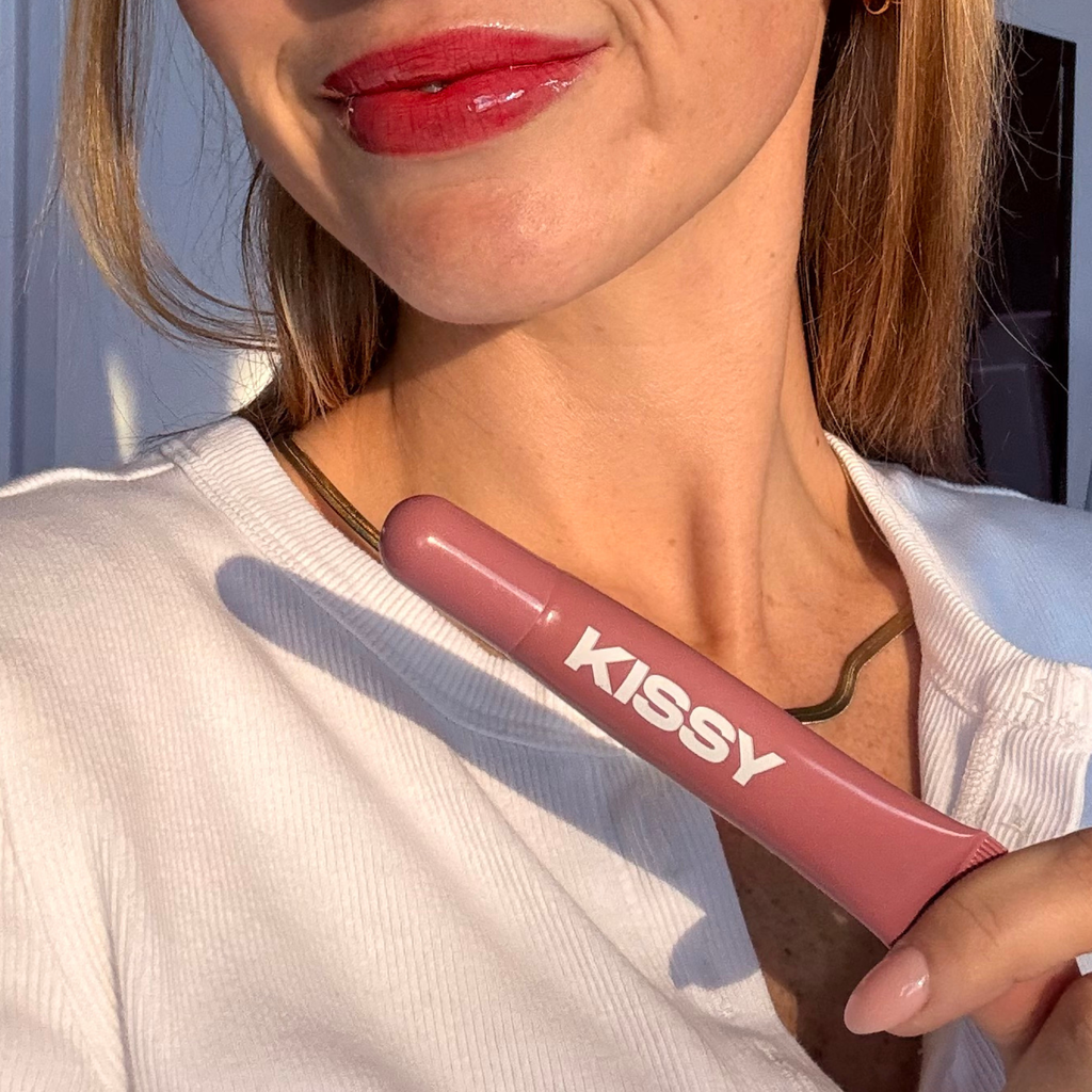 Kissy Gloss Red held beside glossy red lips in sunlight