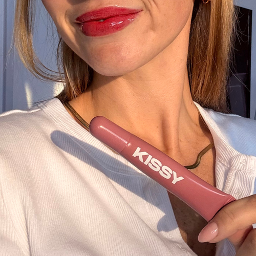 Kissy Gloss Red held beside glossy red lips in sunlight