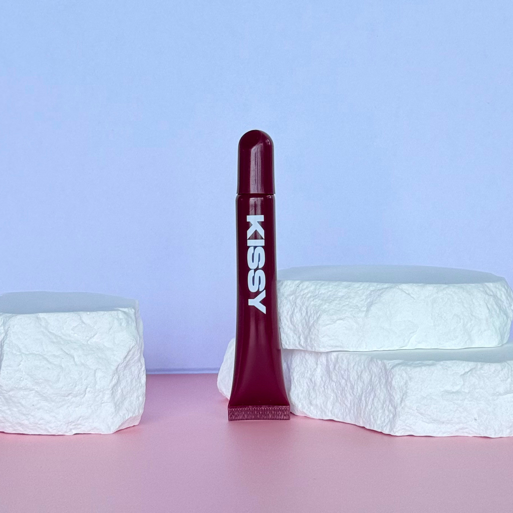 Kissy Gloss Berry held beside glossy berry-toned lips in sunlight